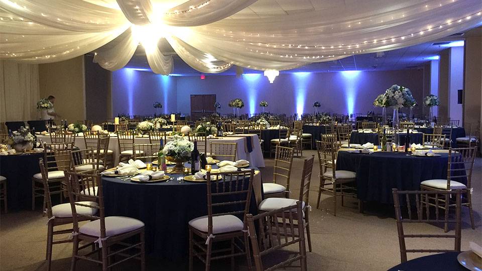 Elegant wedding reception setup in The Pavilion ballroom with navy and gold decor, floral centerpieces, and draped ceiling lights.