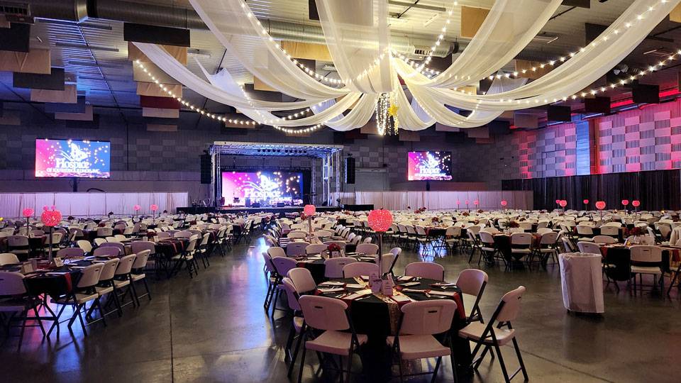 Elegant banquet setup inside The Pavilion Expo Hall with draped ceiling fabric, string lights, and a stage with large display screens.